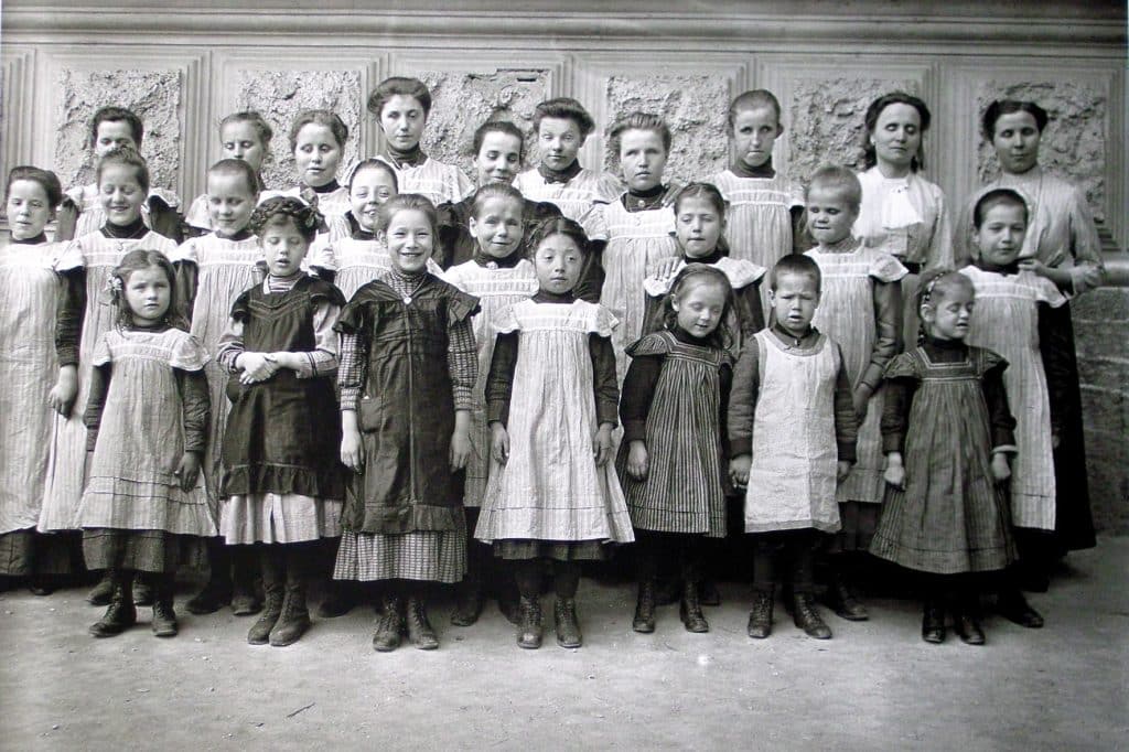 Altes Schwarz-weiß-Foto einer Gruppe Kinder in altertümlichen Kleidern vor einem historischen Gebäude.