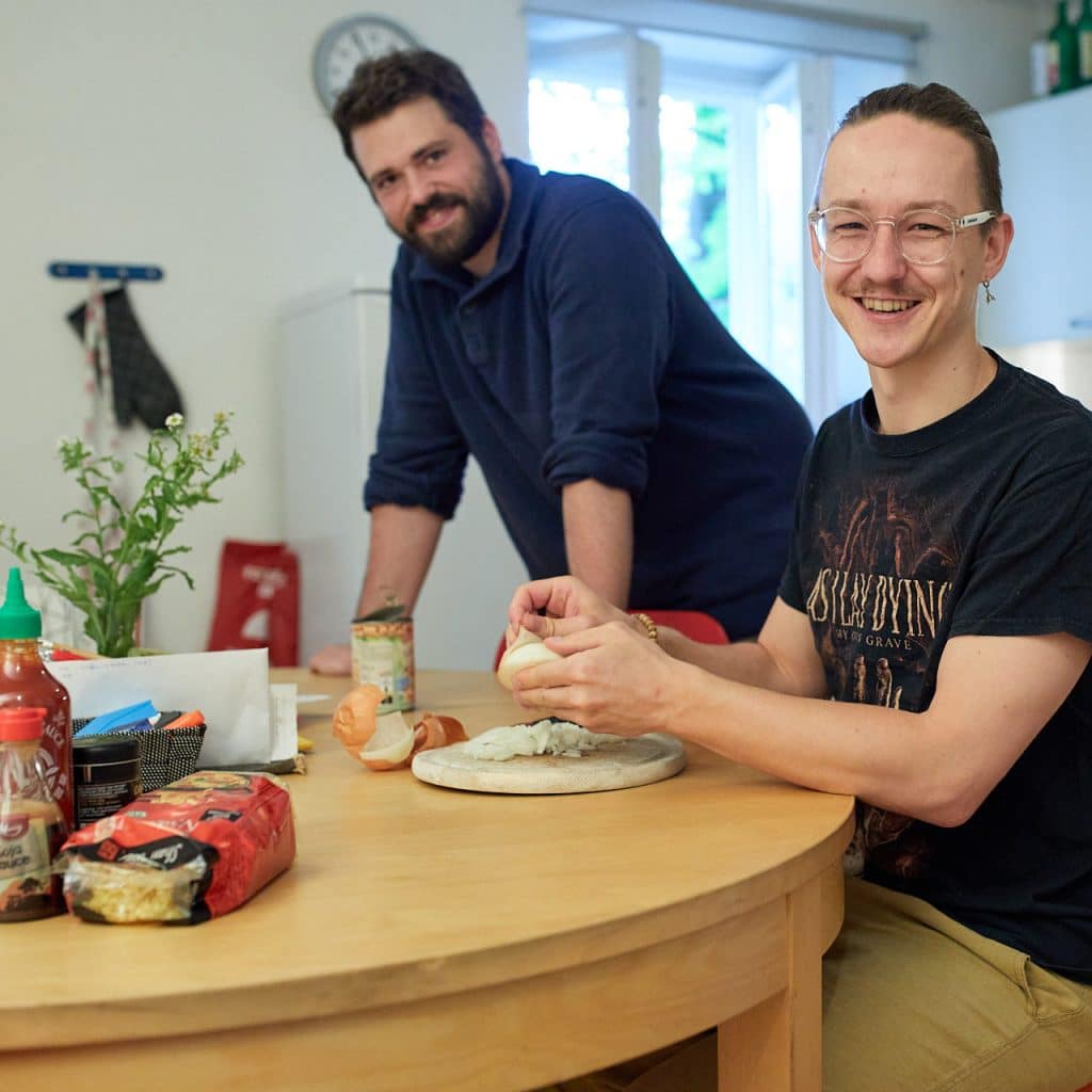 Zwei junge Herren beim Kochen, einer schält eine Zwiebel, beide lächeln.