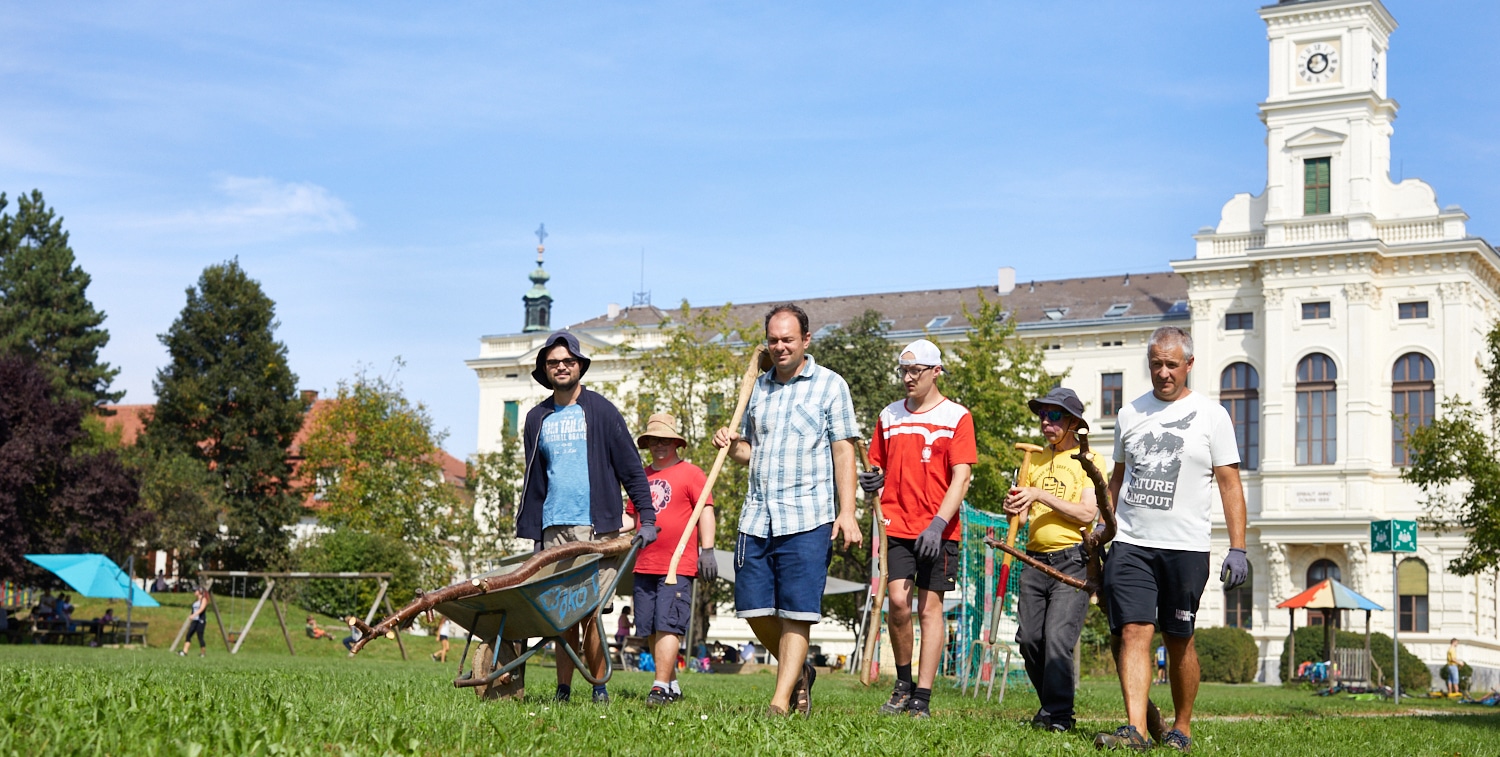 6 Männer gehen mit Gartenwerkzeug und Scheibtruhe vor einem großen, alten Gebäude durch die Wiese.